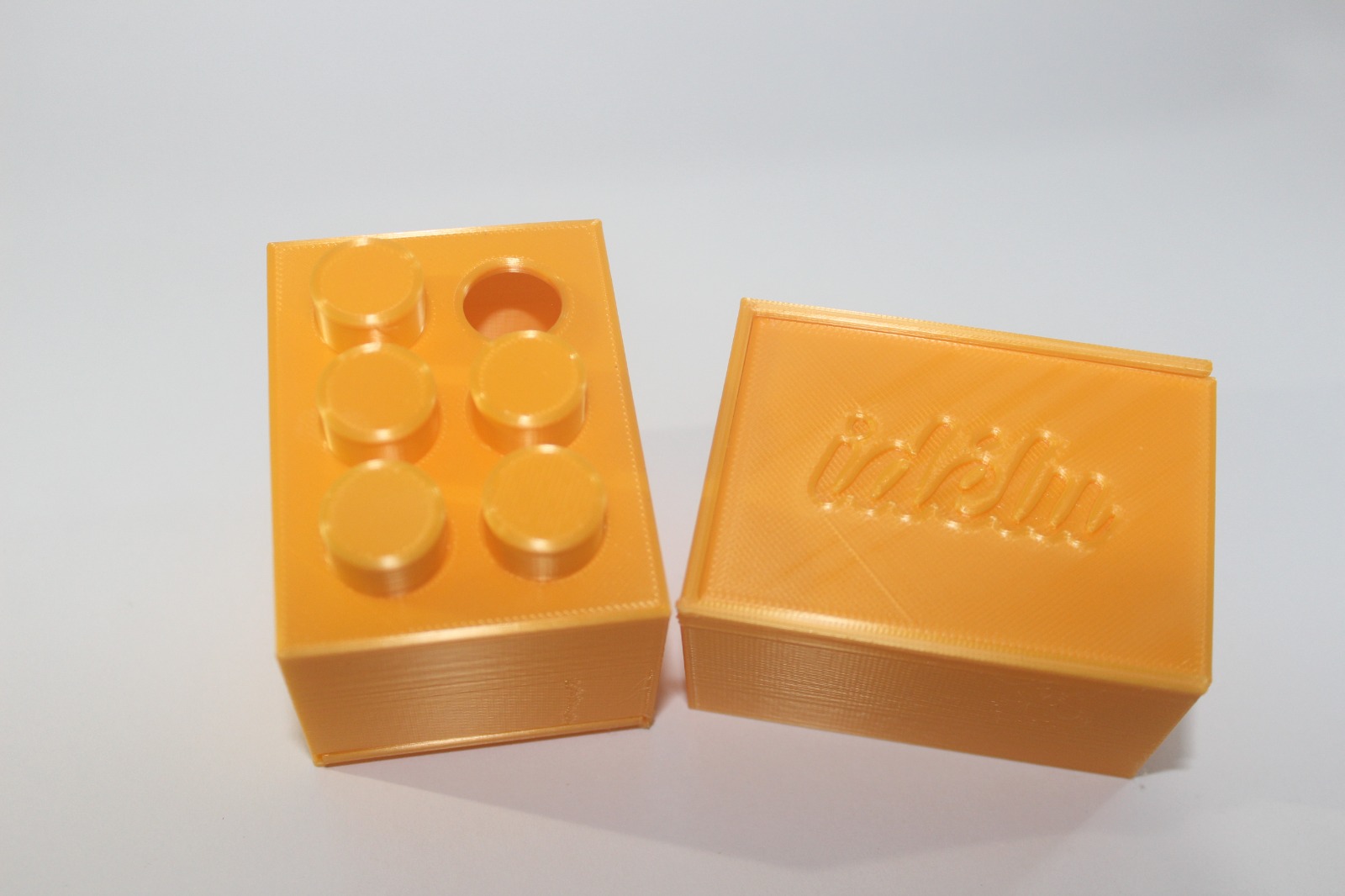 Dos cajas amarillas mostrando perspectivas diferentes del mismo producto. La de la izquierda con una "A" acentuada en Braille hecha con fichas puestas sobre orificios en esa cara de la caja. A la derecha se muestra una cara de la caja con el logo nominativo de Idélu.