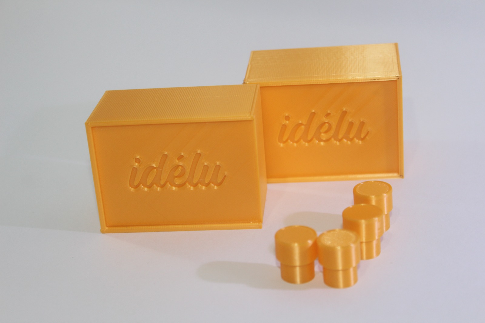 Dos cajas amarillas con el logo nominativo de Idélu y cuatro pequeñas fichas circulares del mismo color al lado, las fichas siendo más delgadas en la parte inferior