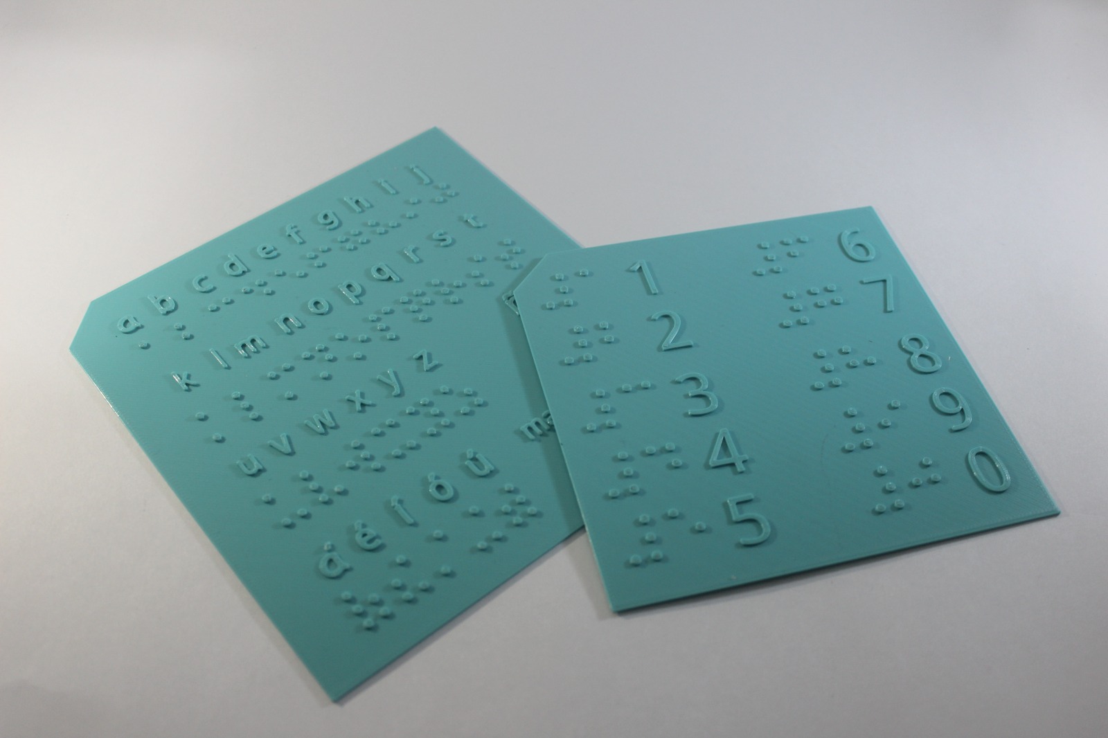 Se muestran dos tablas de color aguamarina con relieve mostrando las letras latinas, en la tabla izquierda, y los números, en la tabla derecha, ambas con su correspondiente en el sistema Braille.